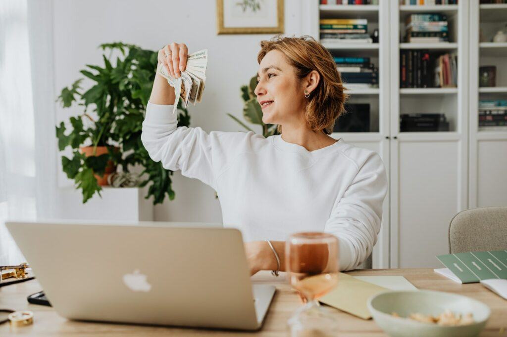 best ways to make money on the side - Lady working at her laptop holding a handful of cash she made on the side